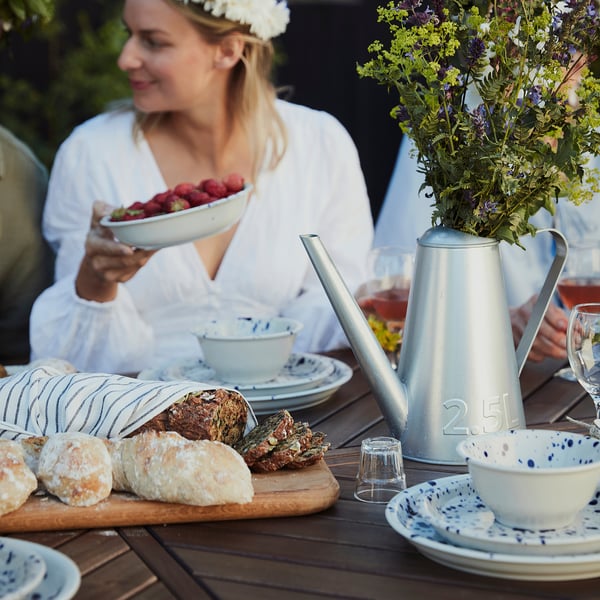 Person holding bowl of strawberries. SILVERSIDA watering can, blue, metal, cylindrical, holds 2. 5l, placed on table with bread and plates.
