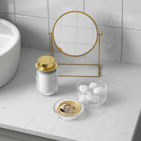 Bathroom vanity set with gold accents: circular mirror, glass jar with lid, and bowls for cotton and jewellery.