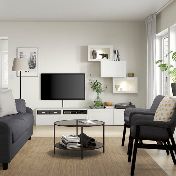 Modern living room with white SINDVIK tv stand, blue sofa, grey chair, and plants. Glass coffee table, large windows.