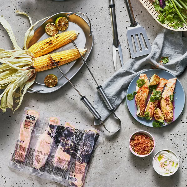Image shows salmon fillets from SJÖRAPPORT, grilled corn, tongs, and seasonings on a grey table.