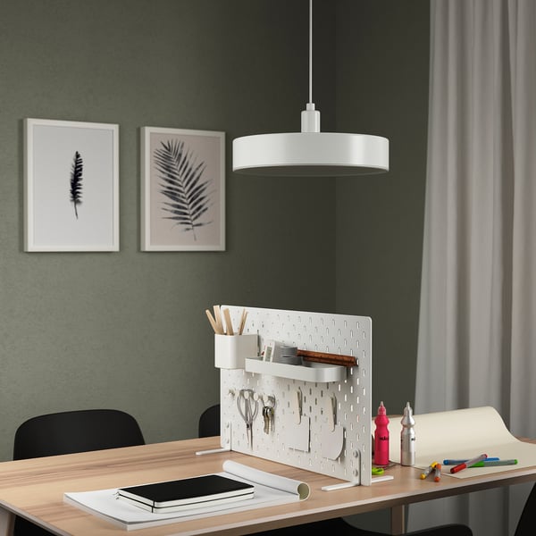 SKÅDIS white desk divider stands tall, organized with office supplies, framed art on wall, lit by hanging lamp.