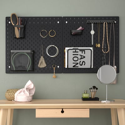 Black SKÅDIS pegboard organiser holds jewellery, makeup tools. Mounted above wooden desk with mirror, makeup items.