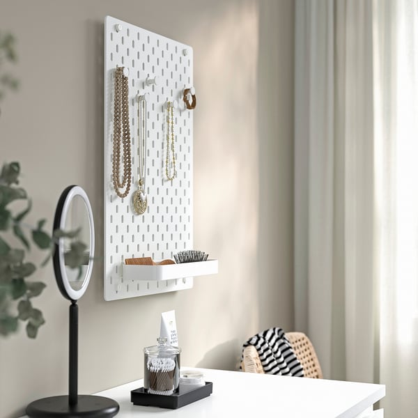 White wall-mounted SKÅDIS pegboard with jewellery and accessories, mirror, and tray on a desk. Organised display and storage.