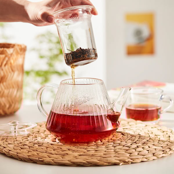 Hand pours tea into glass teapot with strainer on mat near plants.