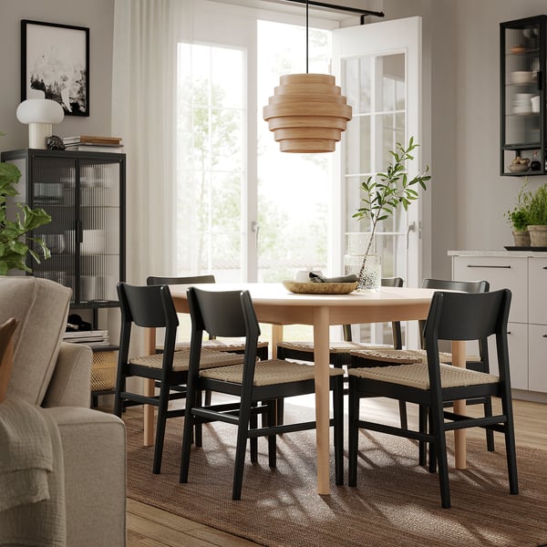 Modern dining room with SKANSNÄS extendable wooden table, surrounded by black chairs with woven seats. Large window and stylish pendant light.