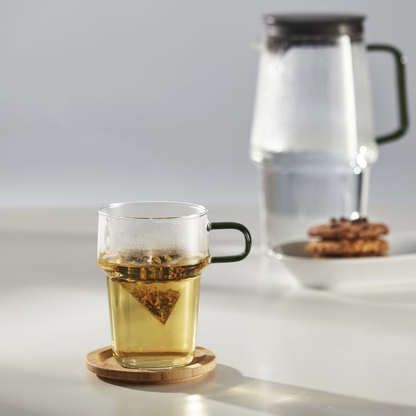 Clear glass mug with green handle holds tea, beside larger pitcher, on white table.