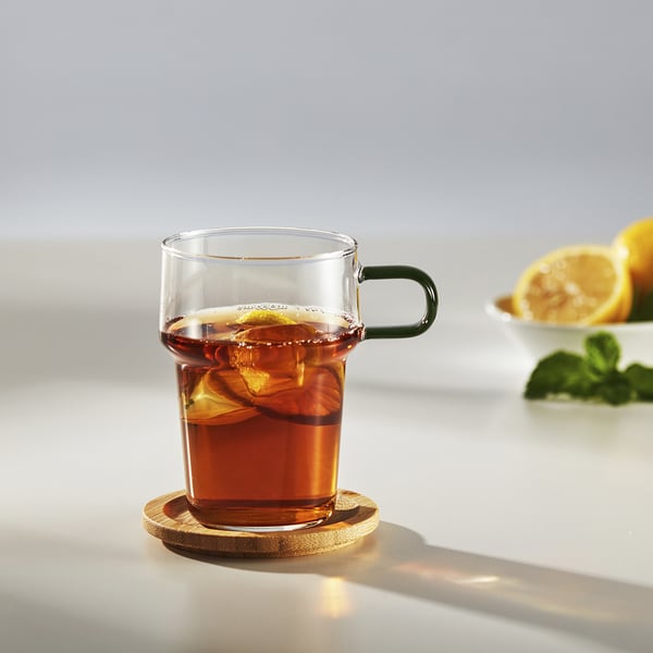 A glass mug with a green handle on a wooden coaster, filled with dark tea and a lemon slice, placed on a light table.