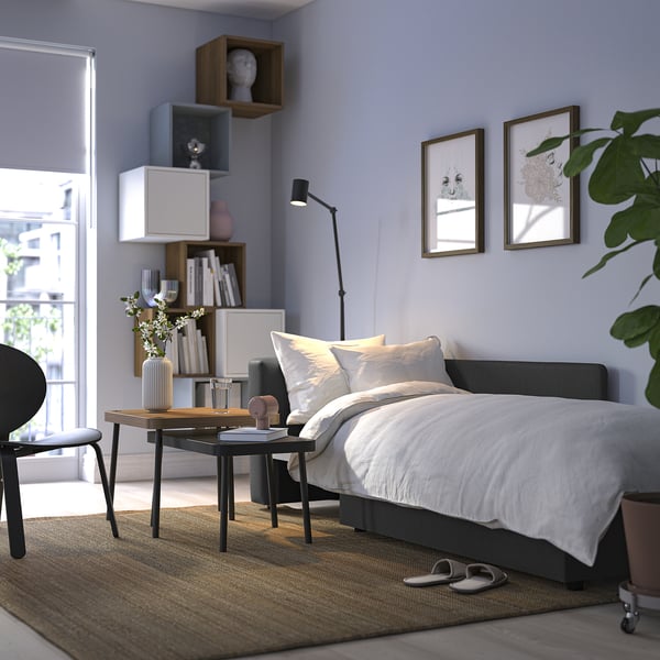 Cosy bedroom with grey sofa bed, white bedding, side table, plants, and decorative shelves.