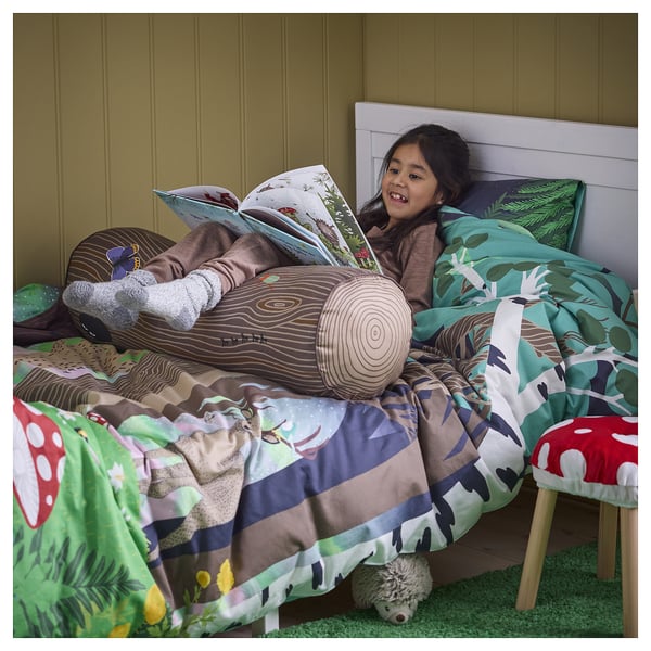 A child on bed with forest-themed pillows, reading. The pillows support their back and head, resembling tree trunks with crawling insects.
