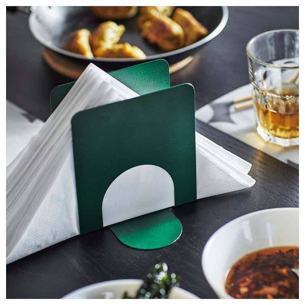 Green SKOGSRÖR napkin holder displays white napkins stylishly on a dark table, with food and drink nearby.