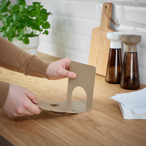Assembling beige napkin holder on table with condiments, plant, cutting board.