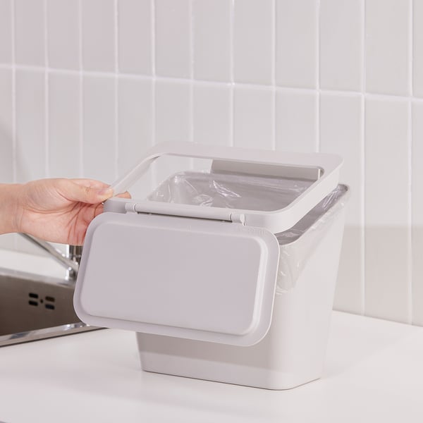 Person opens lid of SKOLÄST waste bin, which hangs on door beside sink. The bin helps keep kitchen tidy and prevents odours.