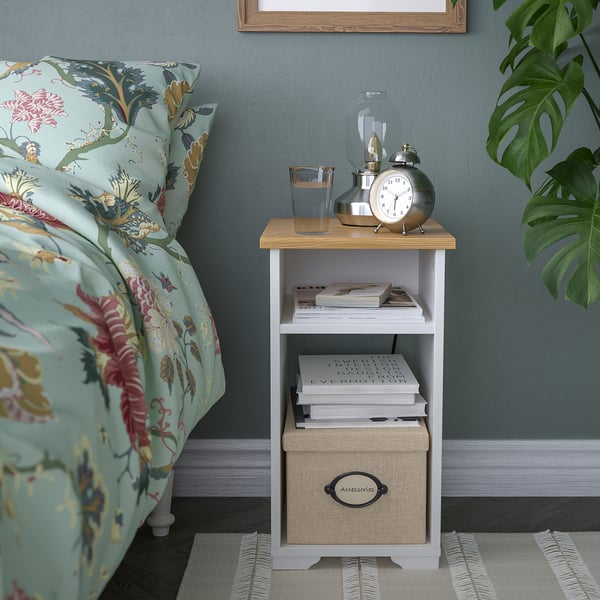 A SKRUVBY side table with a white and wood top, holding books, a clock, and a lamp. Next to a bed with floral bedding.