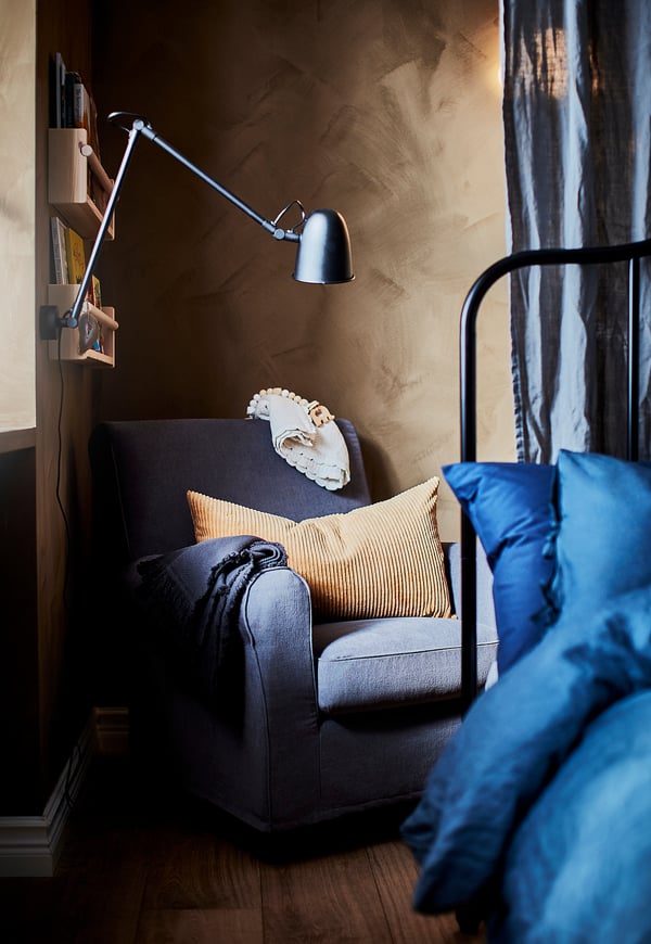 Cosy reading nook with grey SKURUP lamp, arm adjusts, on wall beside blue pillows, grey chair, wooden shelf with books, textured beige wall.