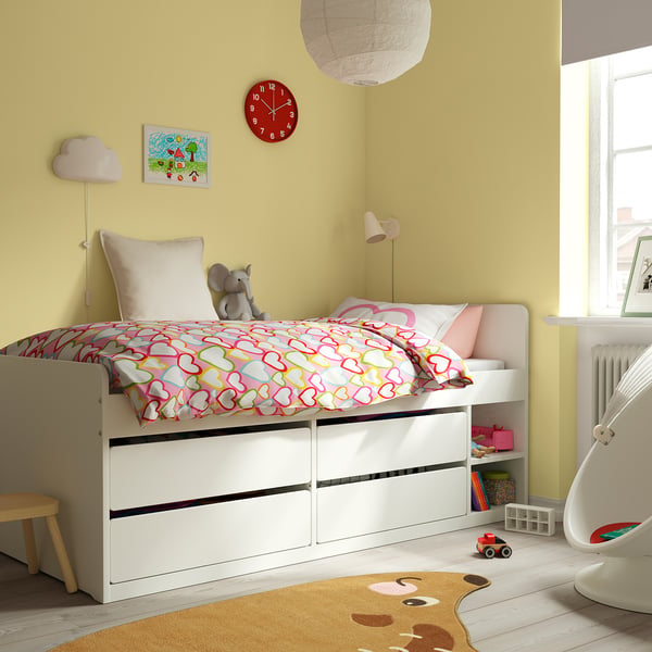 Bright yellow kids room with white SLÄKT storage bed, red clock, two lamps, stuffed elephant, colorful rug, and toys. Bed is central and large.