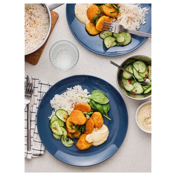 Two blue plates with breaded wheat pieces, rice, cucumber slices, and spinach. Accompanied by bowls of rice and cucumber salad, utensils, and napkins.