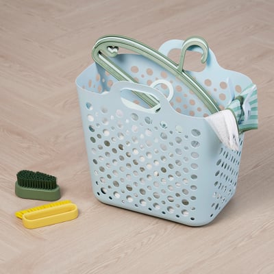 Light blue laundry basket with green handles holds items, two cleaning brushes lie beside it on a wooden floor.