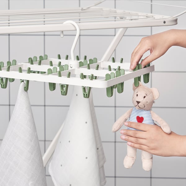 Person hanging white drying rack with green pegs, holding stuffed animal in blue striped outfit with red heart.