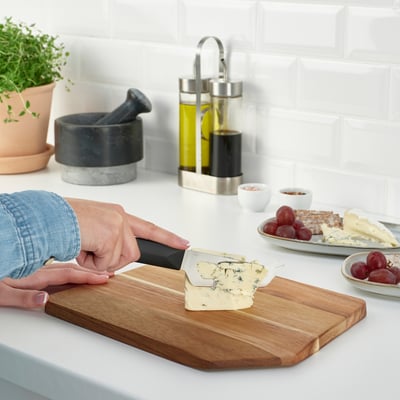 Person slicing cheese on a wooden cutting board on a kitchen counter. Nearby are grapes, crackers, and condiments.