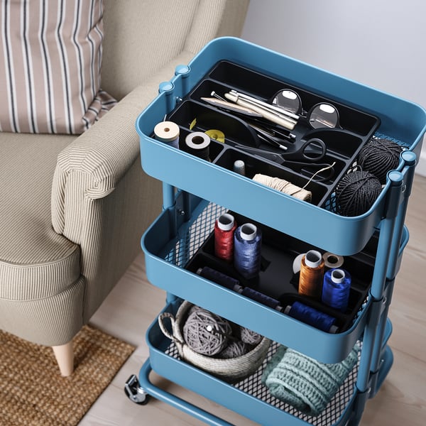 Blue craft cart with wheels, divided trays, holding yarn, tools, scissors, on a rug near striped chair.