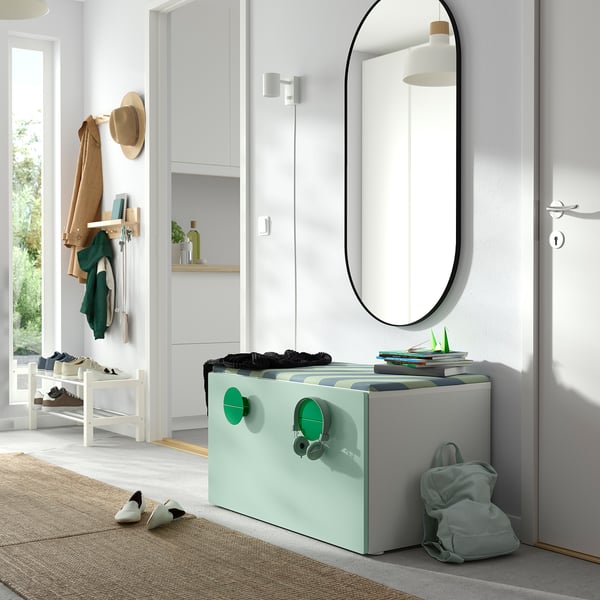Entryway with mint green SMÅSTAD toy box, books, and backpack; white walls, round mirror, coat hooks, and shoe rack.
