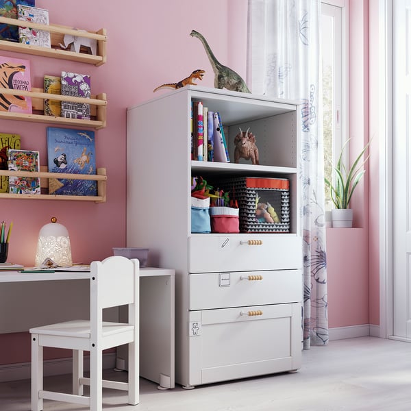 Kids room with SMÅSTAD white storage unit, shelves filled with colourful books, dinosaur toys, and a white chair.