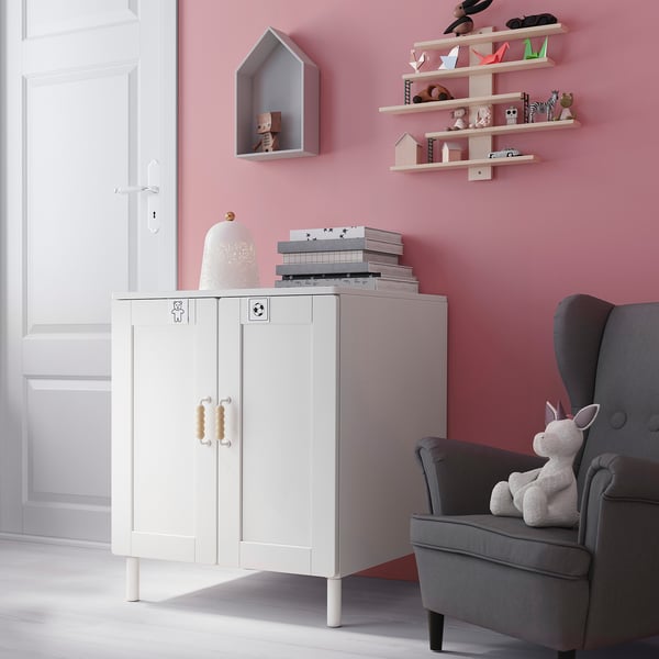 Childs room with pink walls, white cabinet, grey chair, plush unicorn toy, books, and shelves holding toys and decor.