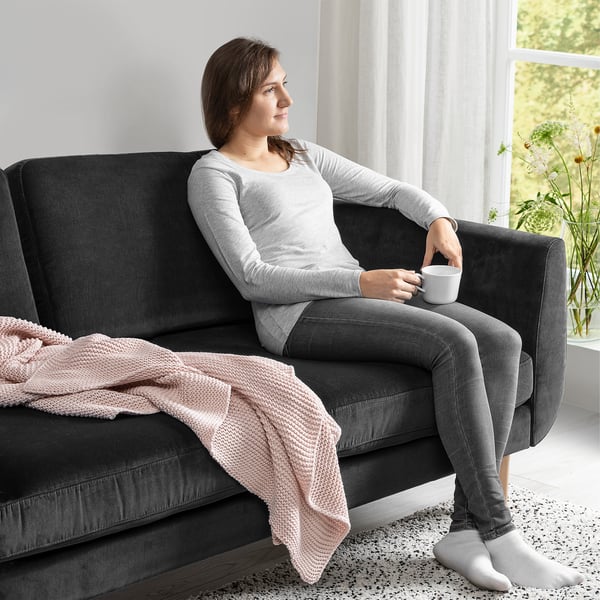 A person sits on a dark, retro-style sofa, holding a mug, with a light pink blanket draped over the armrest.