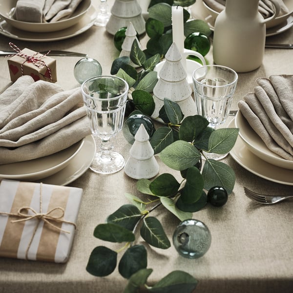Christmas table set with white plates, glasses, grey napkins, and gifts. Centred are SMYCKA garlands with plastic leaves. Small lit trees decorate.
