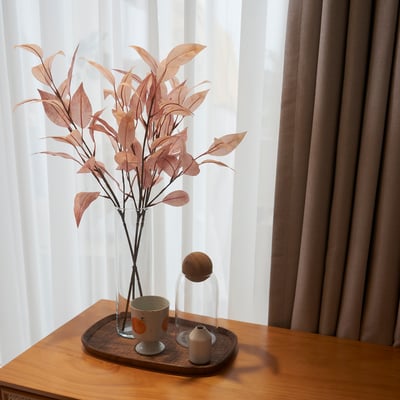 Vase with brown leaves, mug, and candle on wooden table by window.