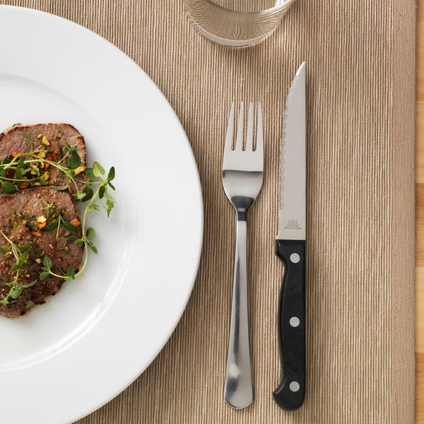 A steak knife with a serrated blade and black handle is placed next to a fork and a plate with cooked meat garnished with greens.