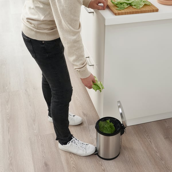 Person discarding lettuce into a stainless steel pedal bin beneath a kitchen counter.