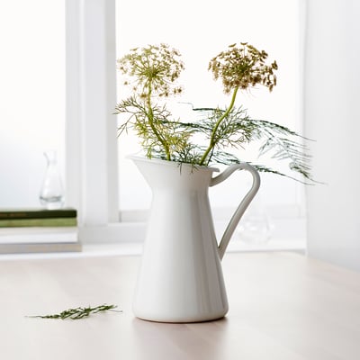 White ceramic jug-shaped vase holding dried flowers, standing on a light-colored surface beside a window, with additional stems on the floor.