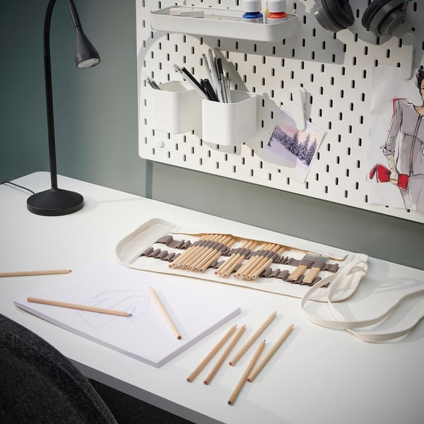 A white desk with SOLFÅGEL colored pencils in a holder; a lamp, sketches, and headphones.