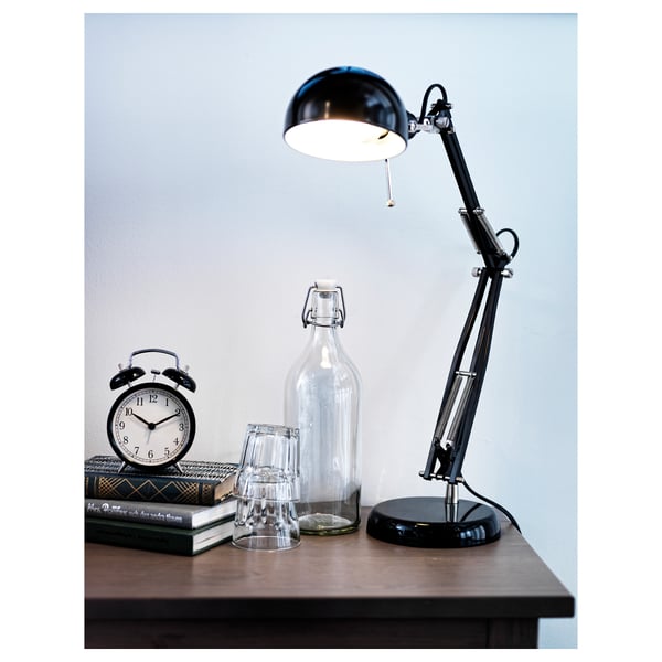 A black desk lamp with a round shade illuminates a wooden desk holding a black alarm clock, two glass bottles, and several books.