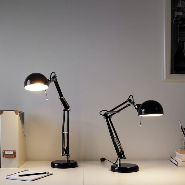 Two black, adjustable SOLHETTA lamps on a white desk. One is smaller, both are lit, positioned near a box and books.