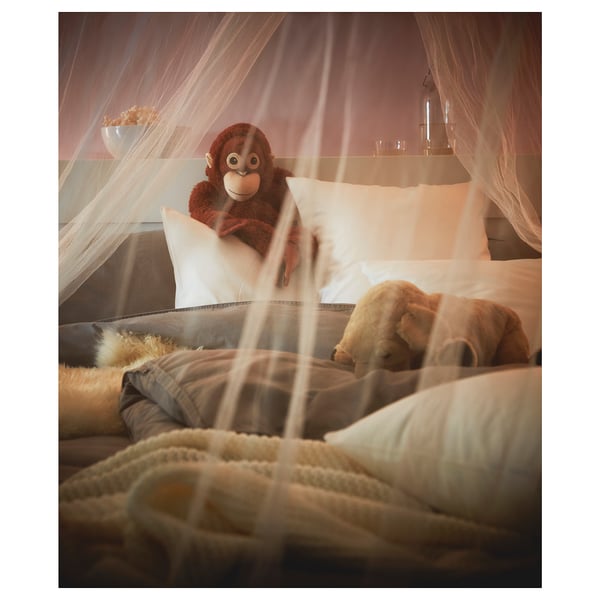 Brown stuffed monkey sits on white bed pillows under sheer white canopy.