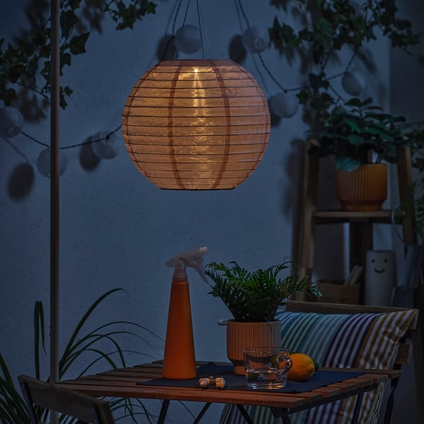 Cosy outdoor setting with a SOLVINDEN solar-powered lantern casting warm light over a table with plants, drinks, and an orange.