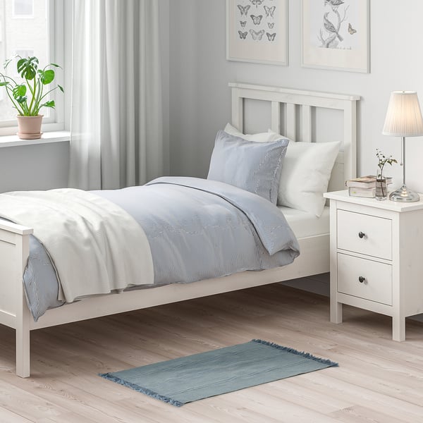 Small SORTSÖ rug in blue, made of cotton, rectangular, with fringe. Positioned near a twin bed, smaller than the bed, adding colour and cosiness.