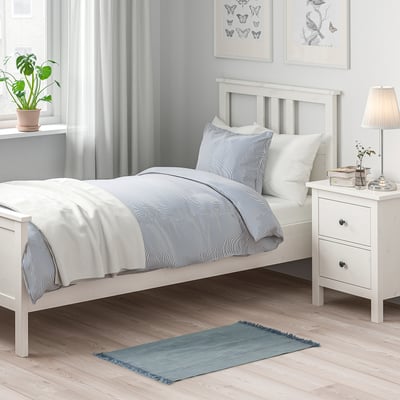 Small SORTSÖ rug in blue, made of cotton, rectangular, with fringe. Positioned near a twin bed, smaller than the bed, adding colour and cosiness.