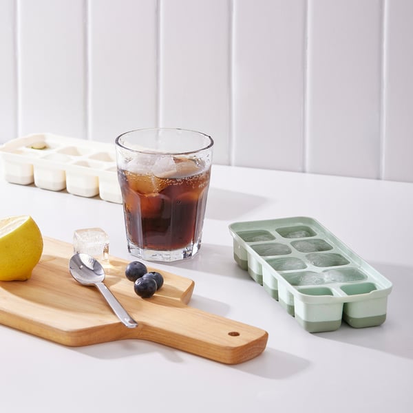 A glass with iced tea sits on a counter next to a tray with frozen fruits and ice cube trays.