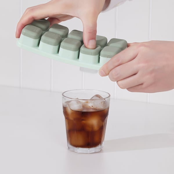 A person uses a spill-proof ice cube tray to fill a glass with ice cubes. The tray has a lid, preventing spills while transferring ice cubes.