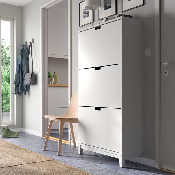 Modern white STÄLL shoe cabinet near grey door, with hanging hooks and a chair.
