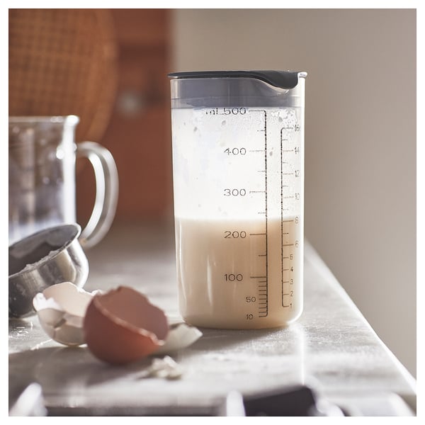 A clear plastic measuring shaker, labelled with ml and oz, stands on a white surface. It has a black lid. An egg shell lies beside it.