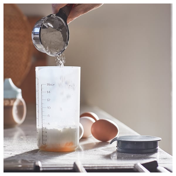 Hand pouring powder into clear measuring cup with black lid.