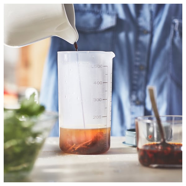 Clear plastic measuring cup with lid pouring amber liquid on white surface.
