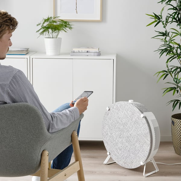 Person sitting in grey chair, using smartphone. White air purifier with handle, on wooden floor beside the person.
