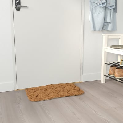 A brown, rectangular STAVREBY doormat made of natural coir, placed near a white door. Its larger than the nearby shoe rack.