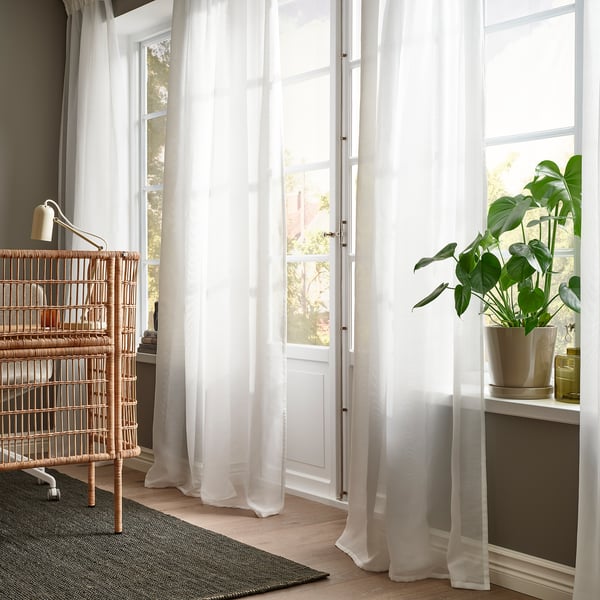 White curtains by tall window soften light, with wicker bookshelf holding books and plant alongside.