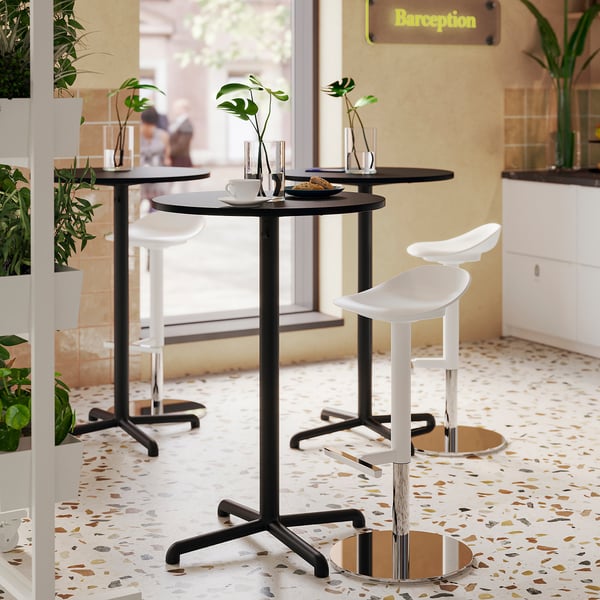 Four black tables with white stools in café. Vases, teacups, bowls. Plants, stone floor. Ideal for dining.